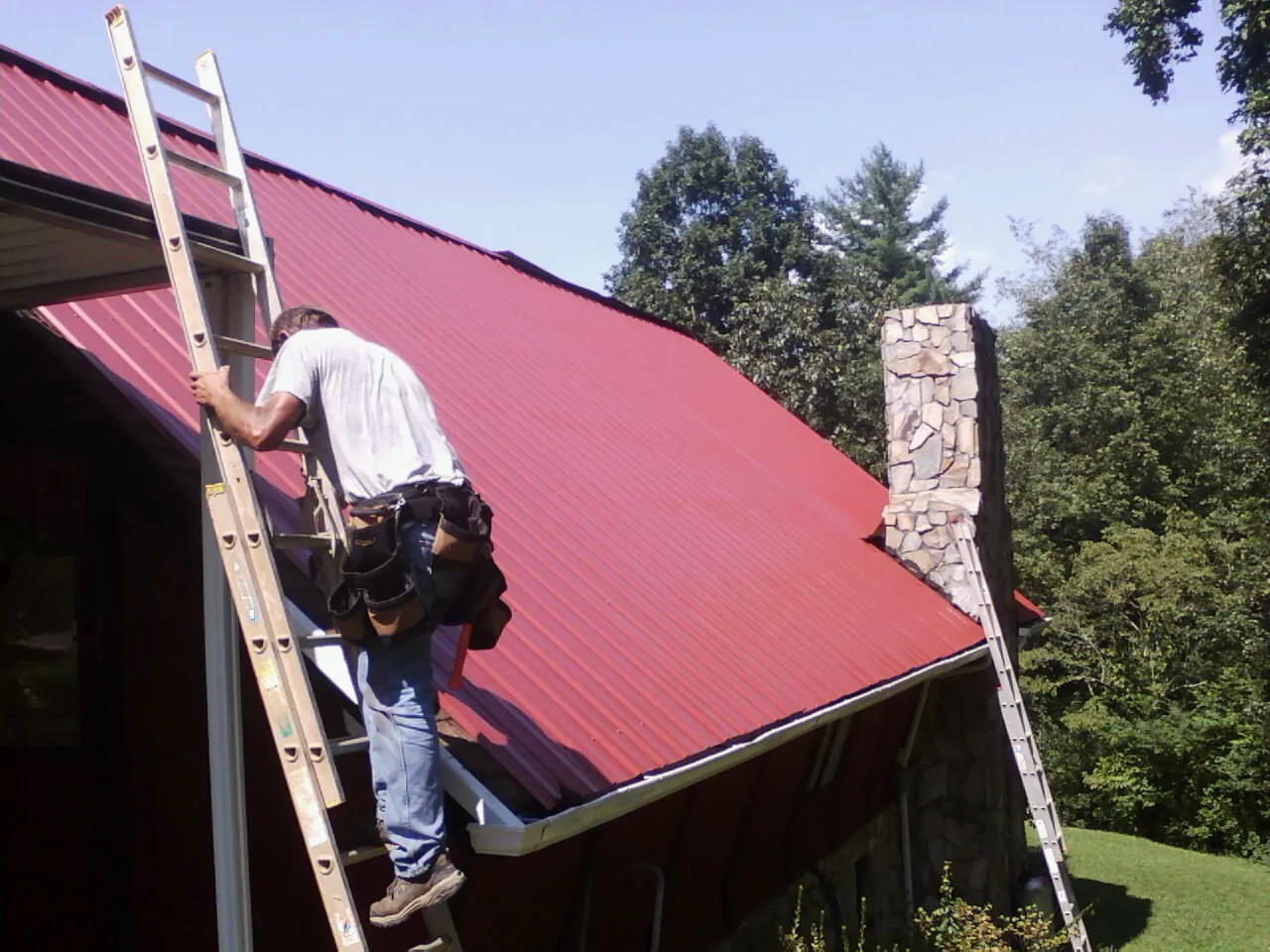 Roofer on a ladder beside a red metal roof and stone chimney — Soffit & Fascia Repair by Roofing Brothers in Bear Creek